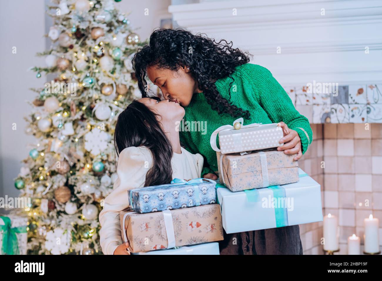 Negro de pelo rizado madre en sombrero de Santa besos en la mejilla hija morena en la diadema de vacaciones sentado contra el árbol de Navidad Foto de stock