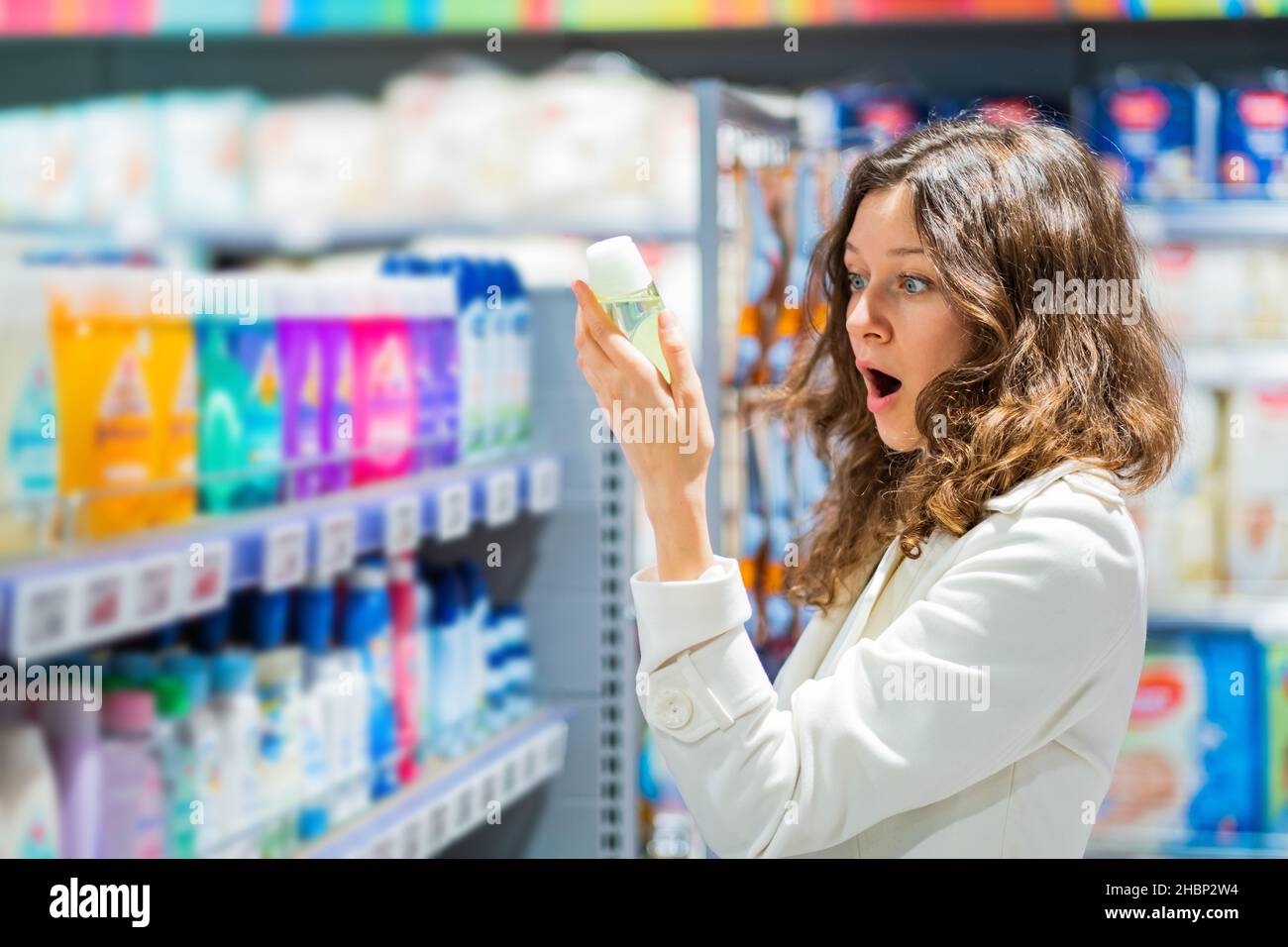mujer en el supermercado leyendo la etiqueta en shock de la composición del champú para bebés, emoción de sorpresa desagradable Foto de stock