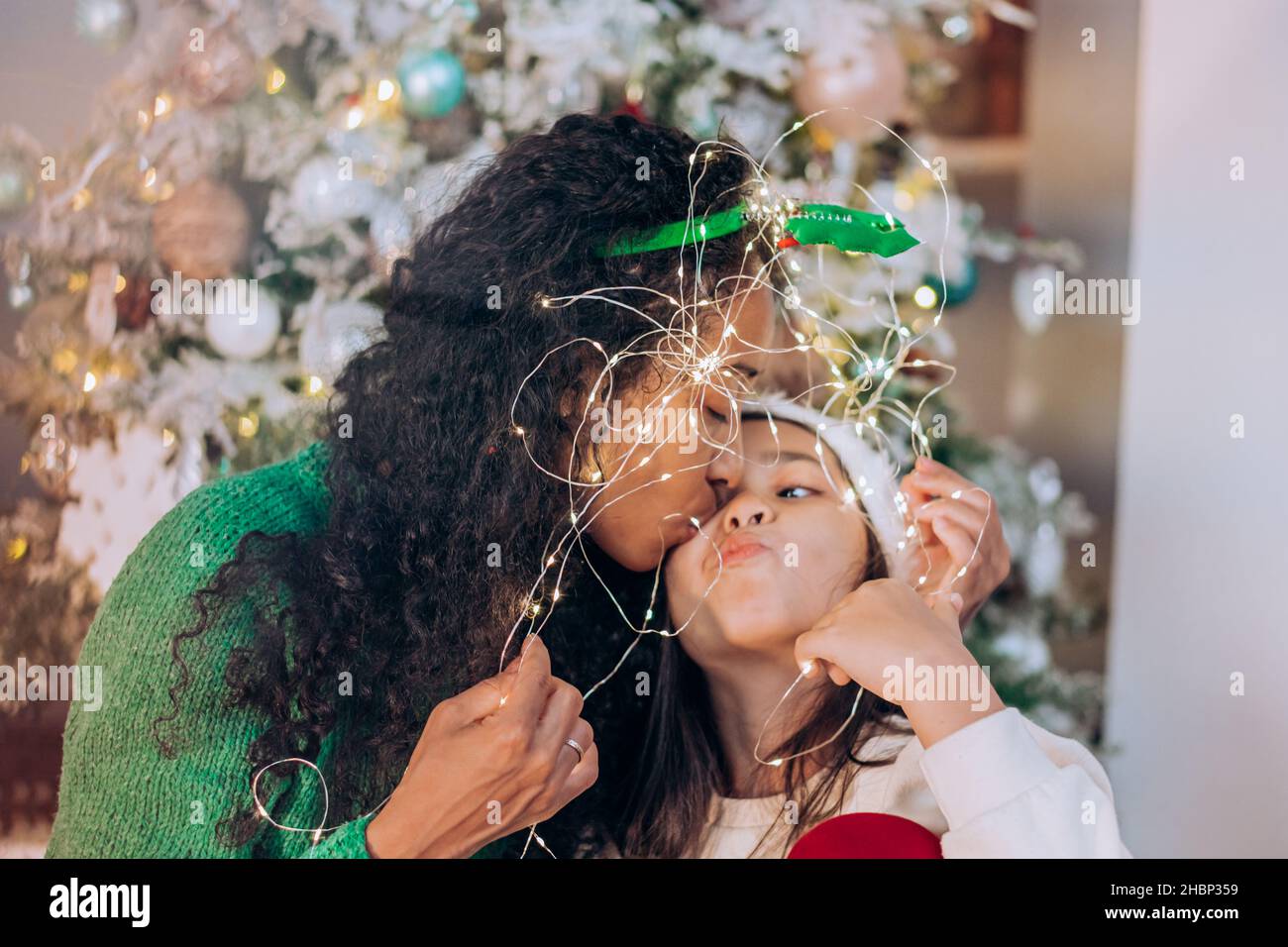 Hija morena con pelo largo y mujer curly afroamericana pose envuelta en guirnalda iluminante contra el árbol de Navidad Foto de stock