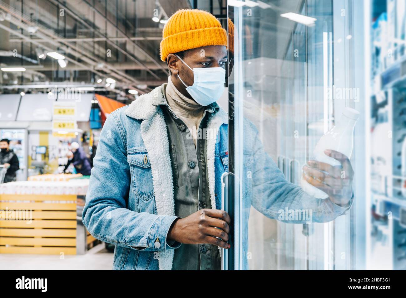 El joven afroamericano con chaqueta vaquera con mascarilla desechable saca la botella de leche de la nevera en el moderno supermercado Foto de stock