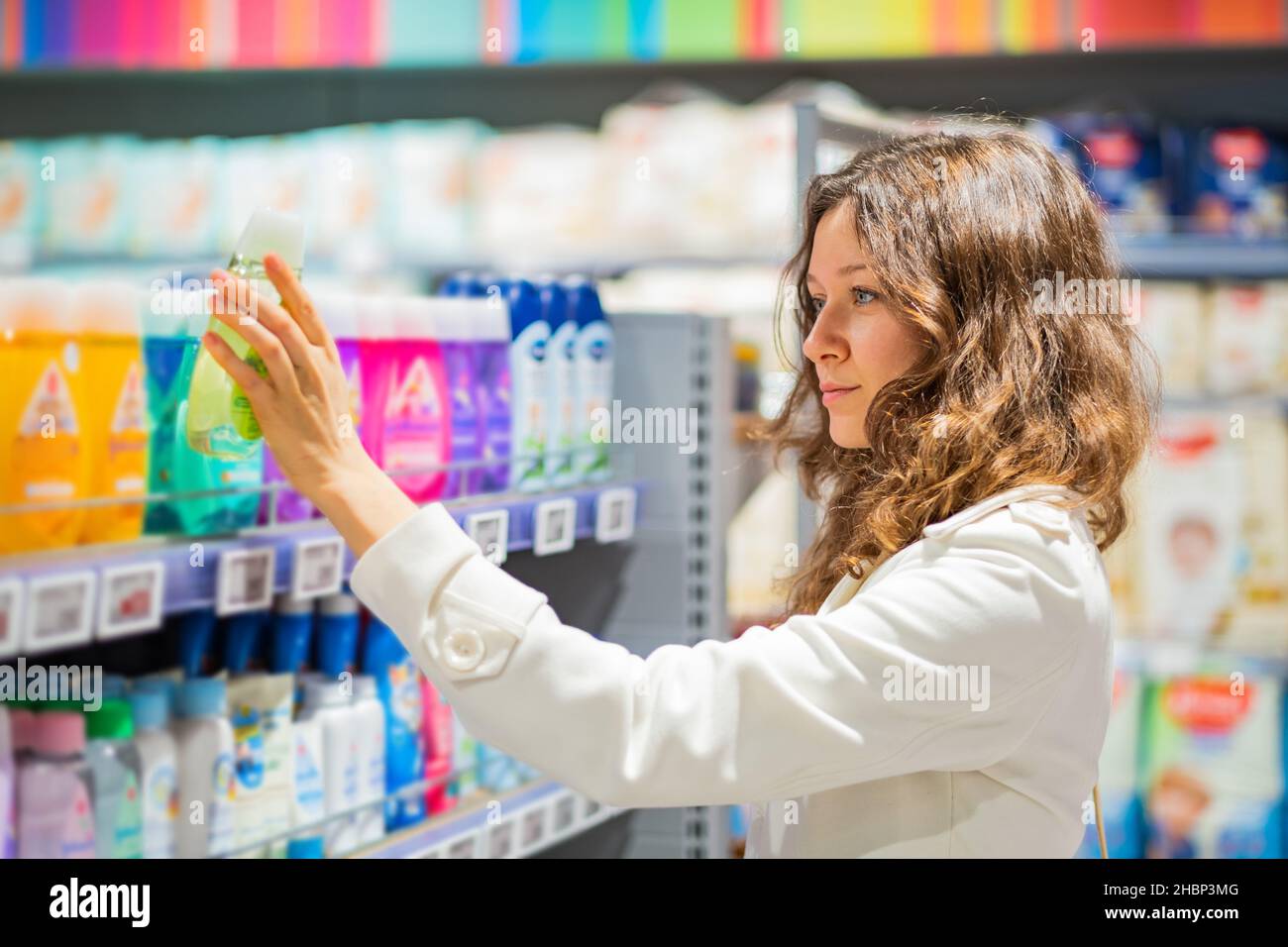 hermosa mujer joven en un abrigo blanco toma champú de la estantería en el supermercado Foto de stock