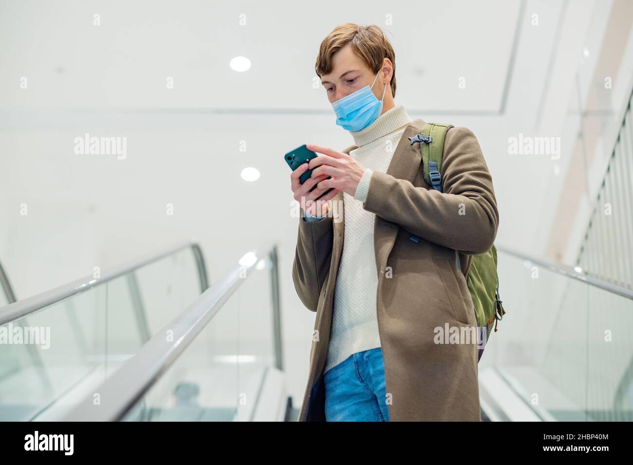 hombre moderno con un abrigo marrón con una mochila y una máscara monta una escalera mecánica en un centro comercial y mira el teléfono en sus manos Foto de stock