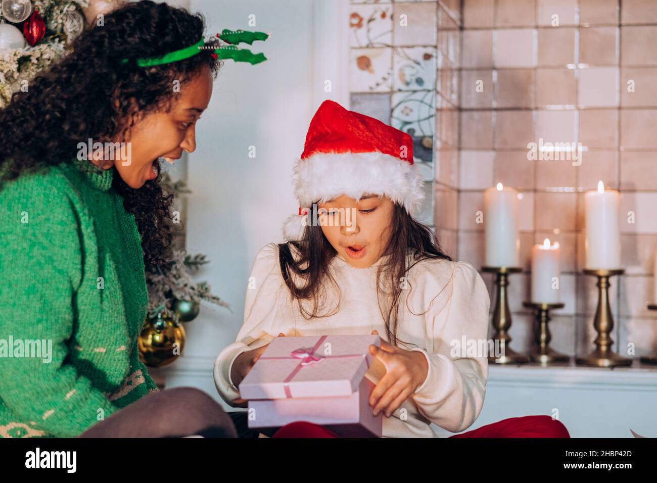 Una niña en un sombrero rojo abre un regalo de Navidad de mamá contra el fondo de un árbol de Navidad y velas, mamá se sienta cerca y espera a su hija para abrir el regalo Foto de stock