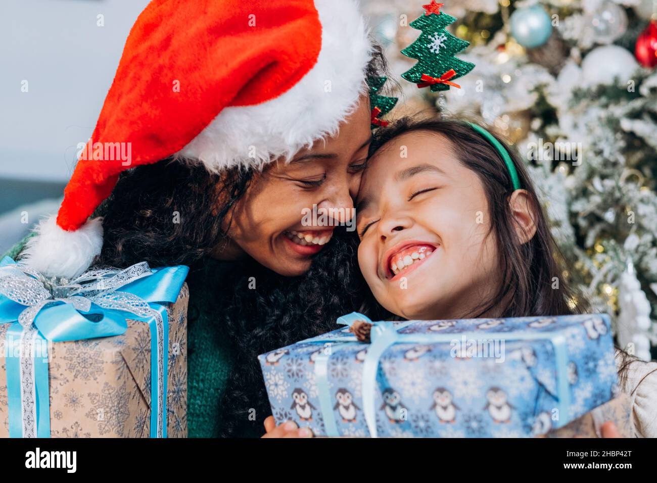 Mamá e hija en una gorra roja están llevando a cabo regalos de Navidad y riendo contra el fondo de un árbol de Navidad, cerca de arriba Foto de stock