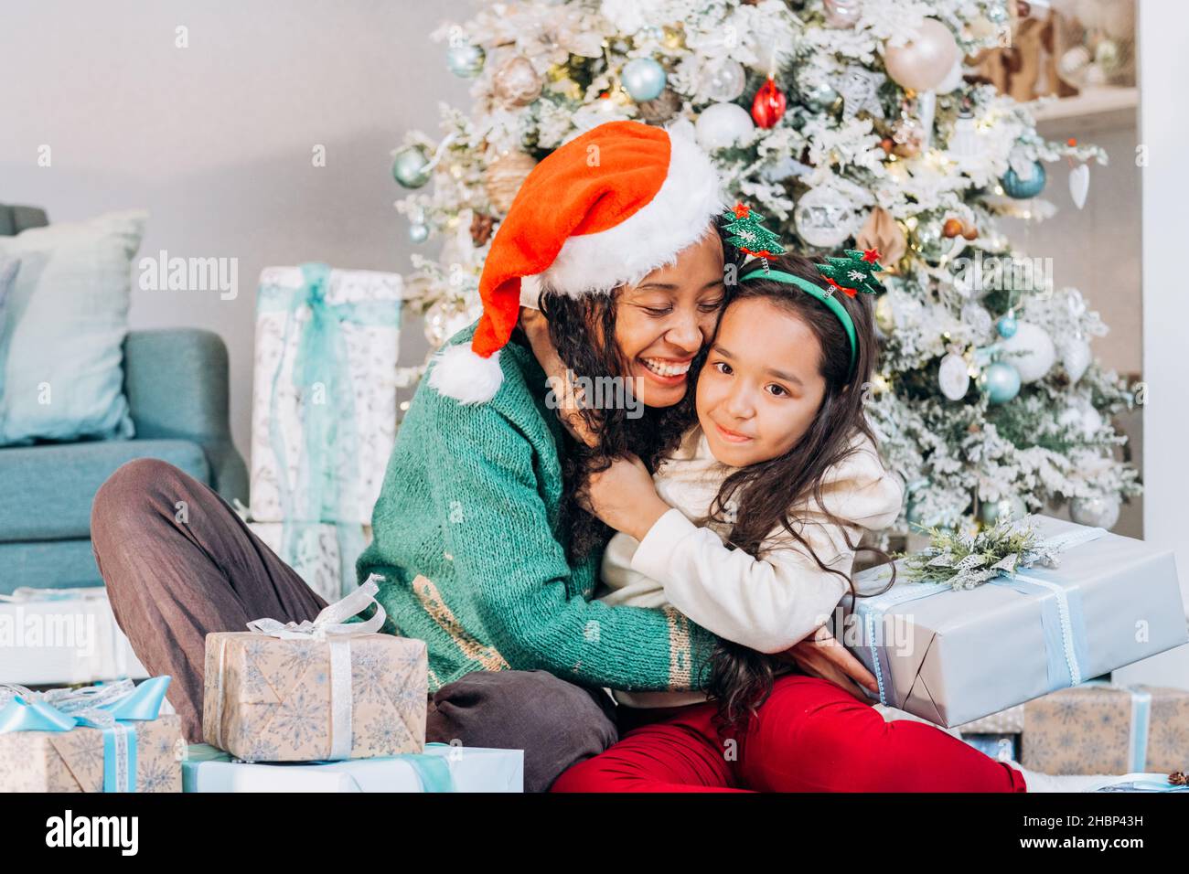 La madre afroamericana y la hija morena de pelo largo en sombreros festivos abrazan fuertemente sonriente sentado entre cajas de regalo por el árbol de Navidad Foto de stock