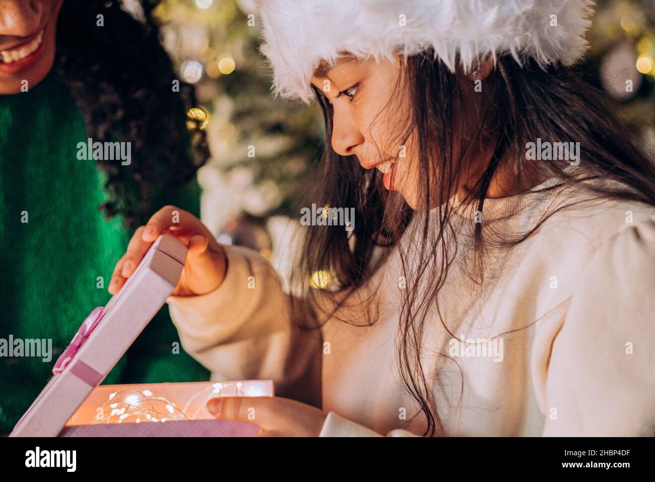 Pequeña niña en un sombrero de Navidad abre una caja con luces brillantes y un regalo en el fondo de un árbol de Navidad junto a su madre Foto de stock