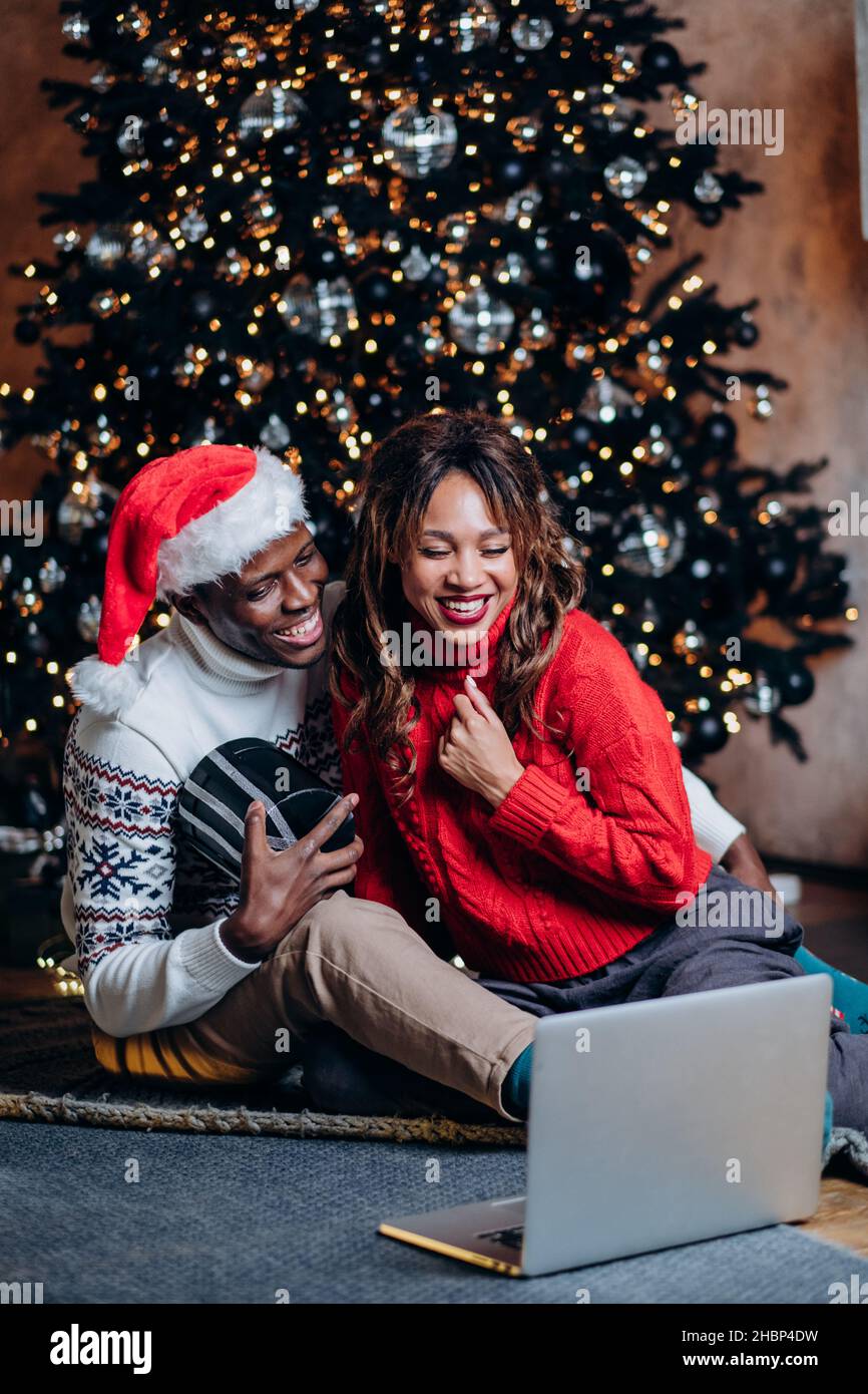 feliz pareja afroamericana felicitando y haciendo videollamadas en el portátil, sentado en el suelo con un regalo contra el fondo de un árbol de navidad, las nuevas tecnologías foto vertical Foto de stock