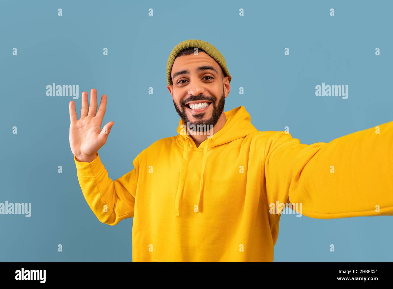 Hola. Feliz hombre árabe tomando selfie y moviendo la mano a la cámara, gesturing hola mientras que hace la foto sobre fondo azul Foto de stock