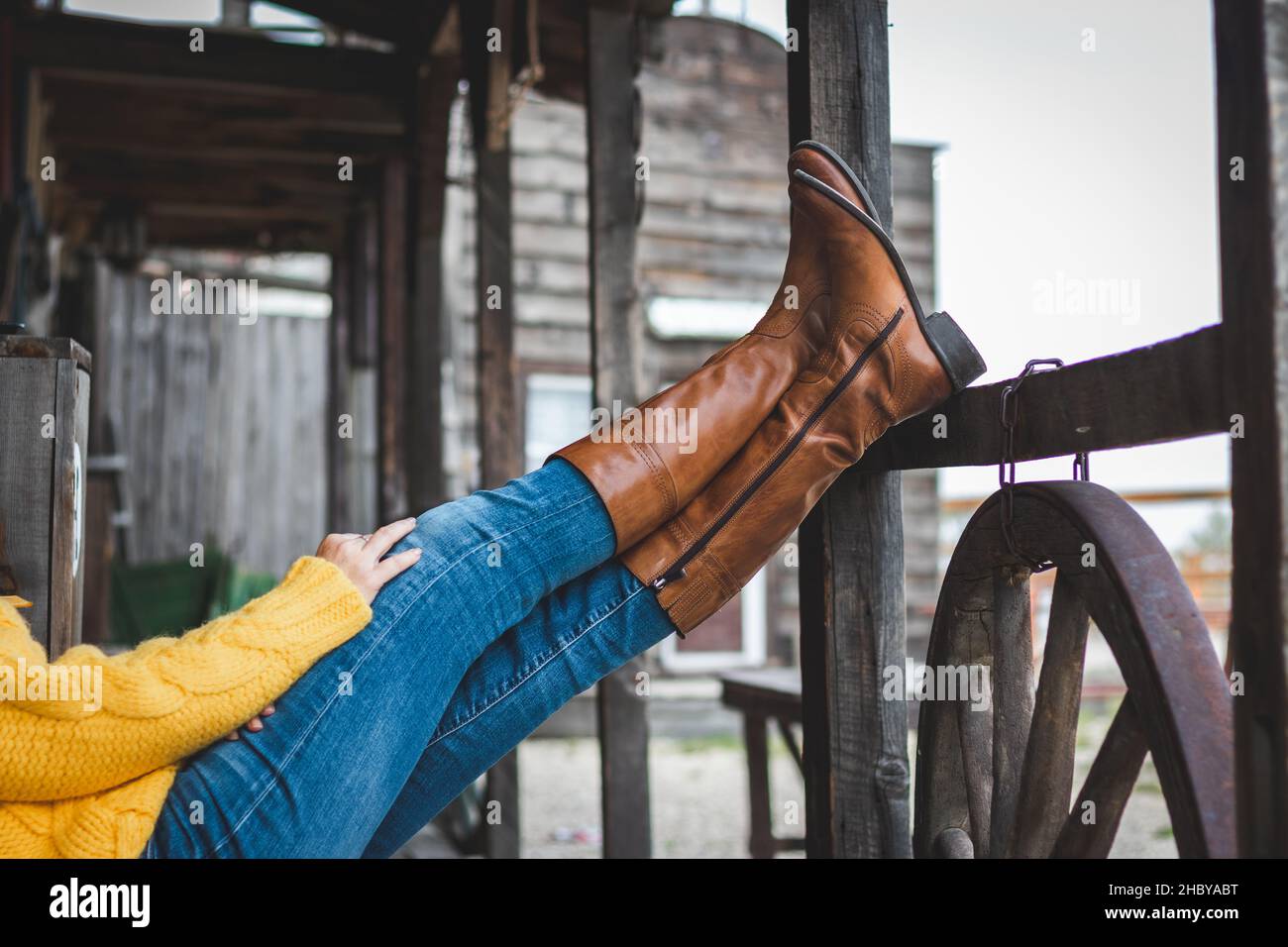 La mujer está relajándose en el rancho con sus piernas en una barandilla de madera. Botas y jeans de cuero Foto de stock