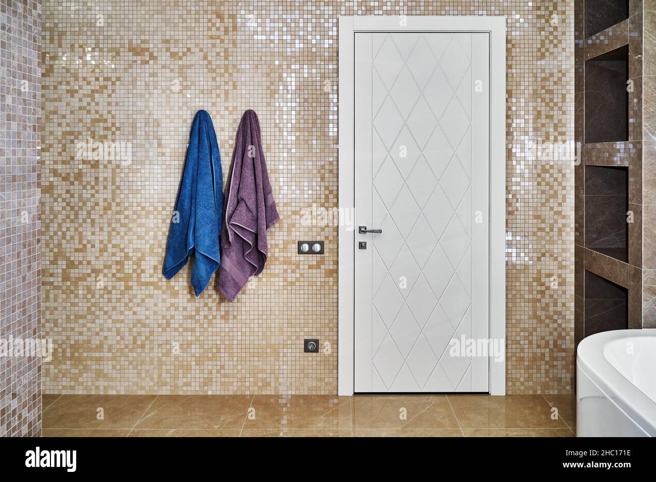Elegante puerta blanca cerrada con diseño de rombos y toallas de felpa colgantes en un amplio baño luminoso decorado con azulejos de cerámica con estampado beige Foto de stock