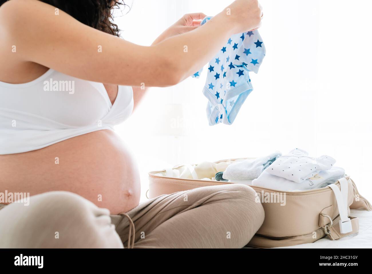 mujer embarazada joven preparando la maleta del bebé para el hospital doblando y poniendo la ropa pequeña para su recién nacido Foto de stock