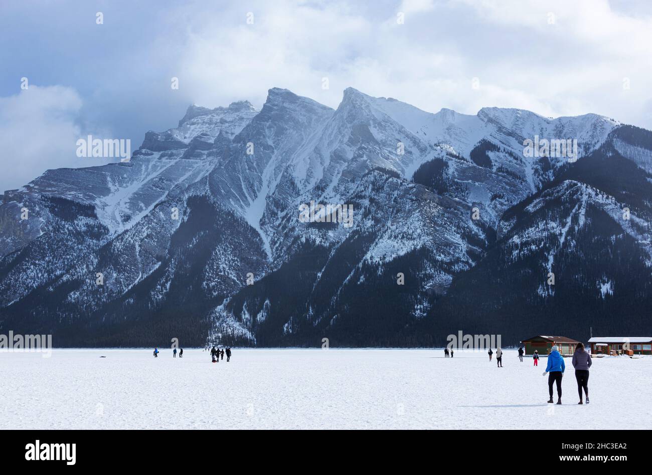 Visitantes inidentificables caminando por el lago congelado Minnewanka en las Montañas Rocosas Canadienses del Parque Nacional Banff durante el frío invierno con el Monte Inglismaldi Foto de stock