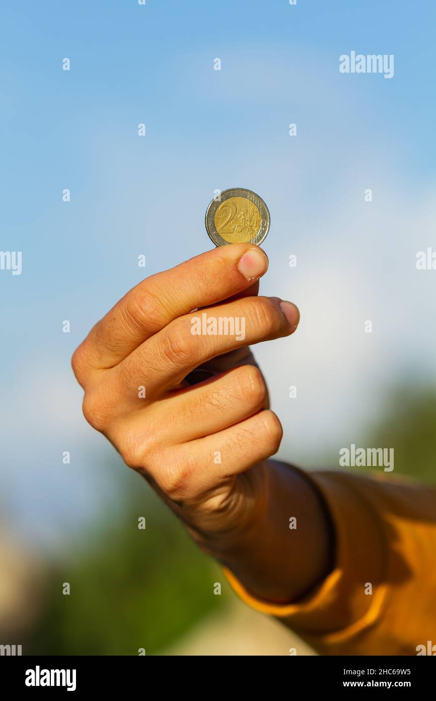 Un plano vertical de la mano de un niño sosteniendo una moneda de dos euros sobre un fondo azul del cielo Foto de stock