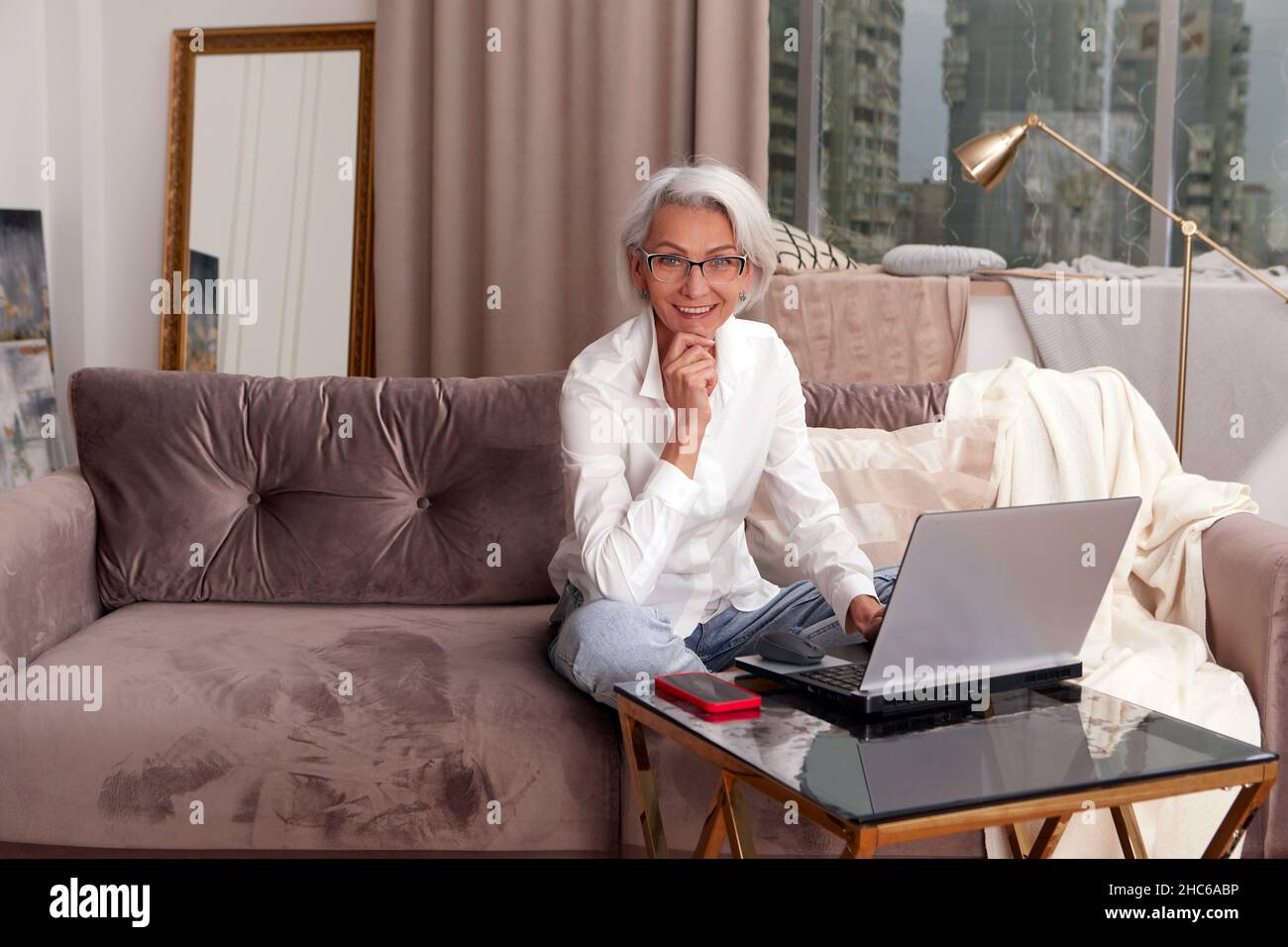 Mujer positiva con el pelo corto en la ropa formal sentado en un sillón suave y mirando la cámara mientras que hojea el portátil en la habitación ligera Foto de stock