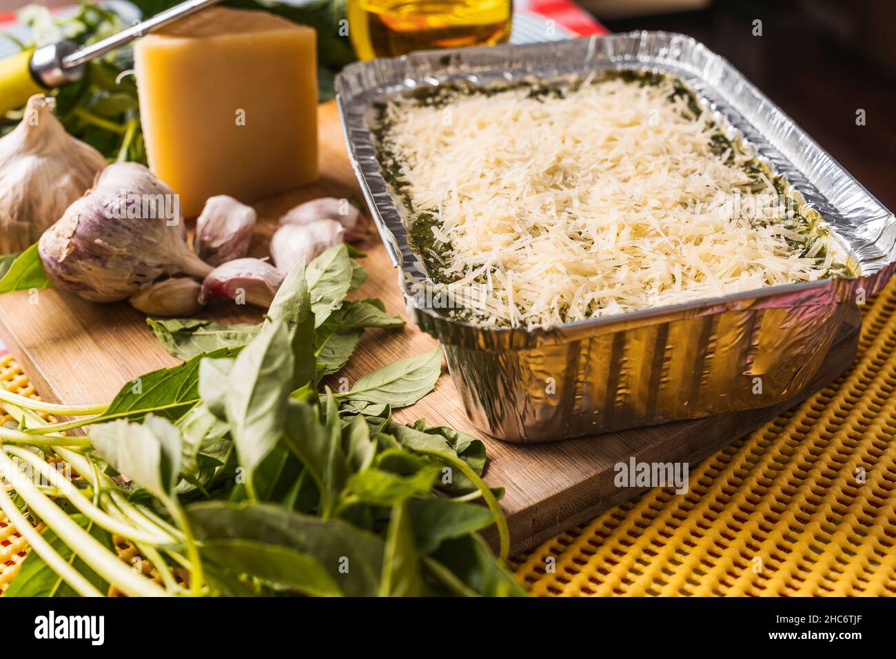 Lasaña casera con pesto en una bandeja de aluminio con un montón de queso rallado la parte superior una tabla de madera y los diferentes ingredientes crudos que componen Fotografía