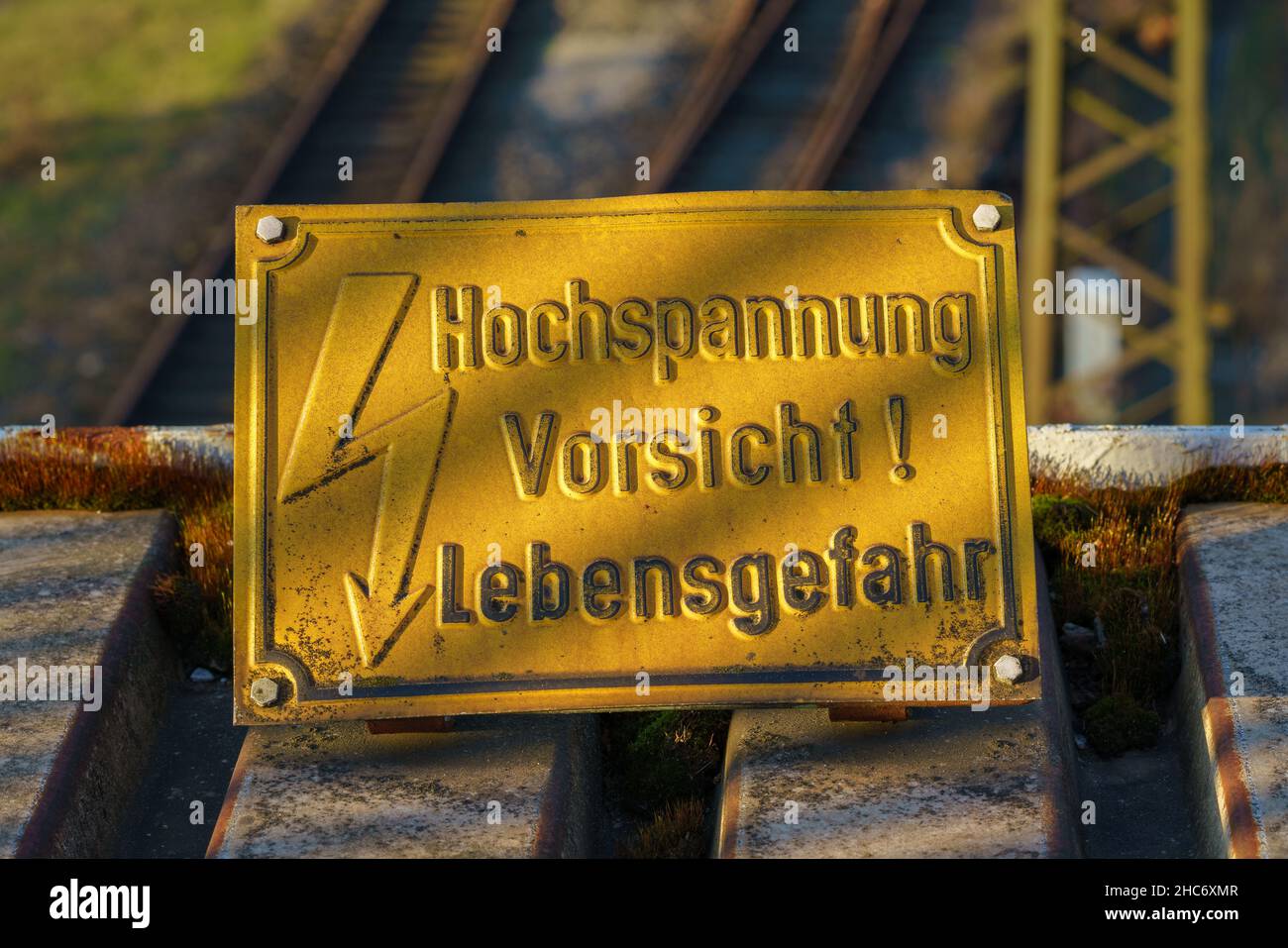 Señal: Alta tensión, peligro de muerte (en alemán: Hochspannung, Vosicht Lebensgefahr), con vías de ferrocarril en el fondo borroso Foto de stock