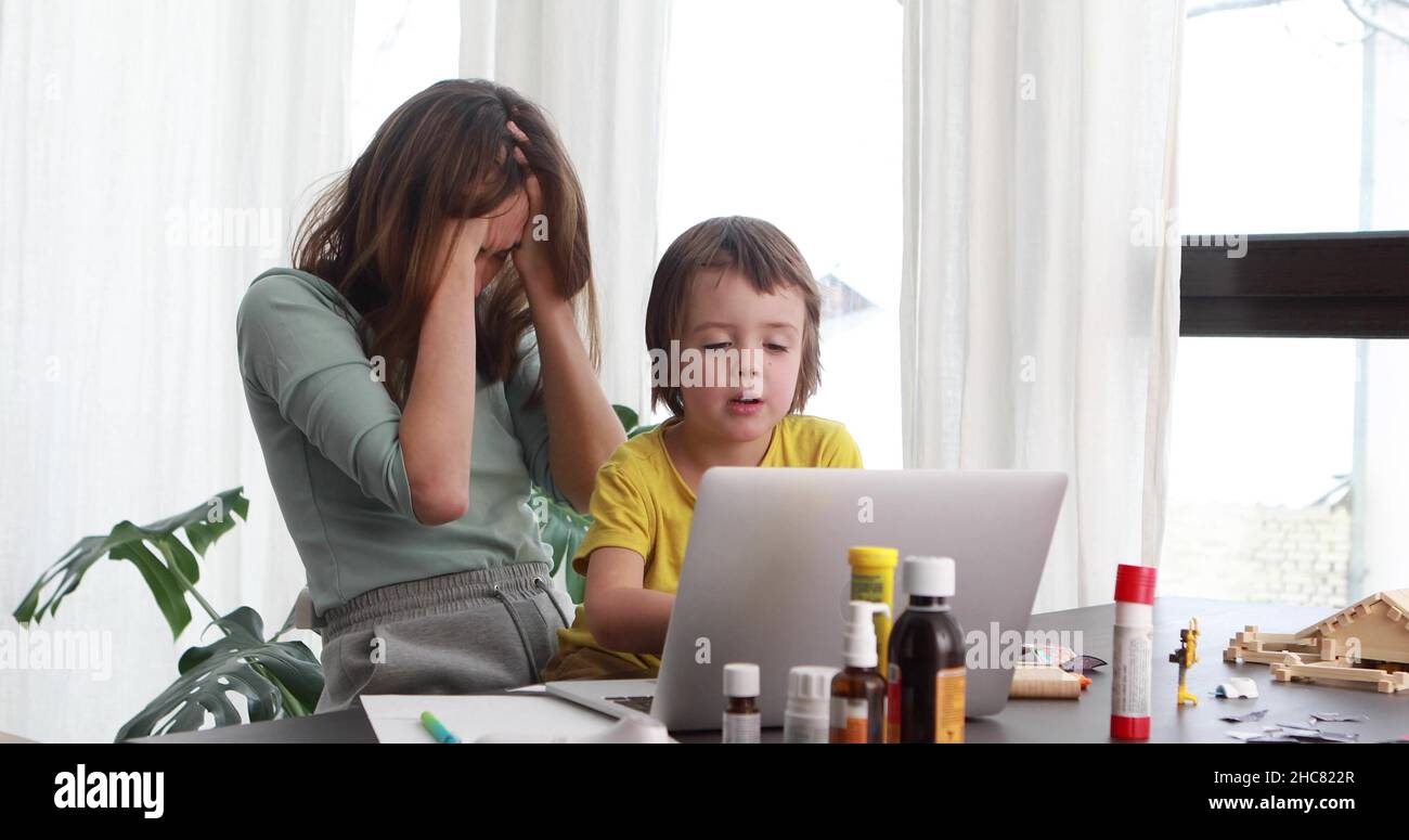 Una mujer cansada se agarra la cabeza mientras trabaja en casa, mientras que un niño pequeño presiona las teclas del teclado del portátil en la mesa con botellas de farmacia Foto de stock