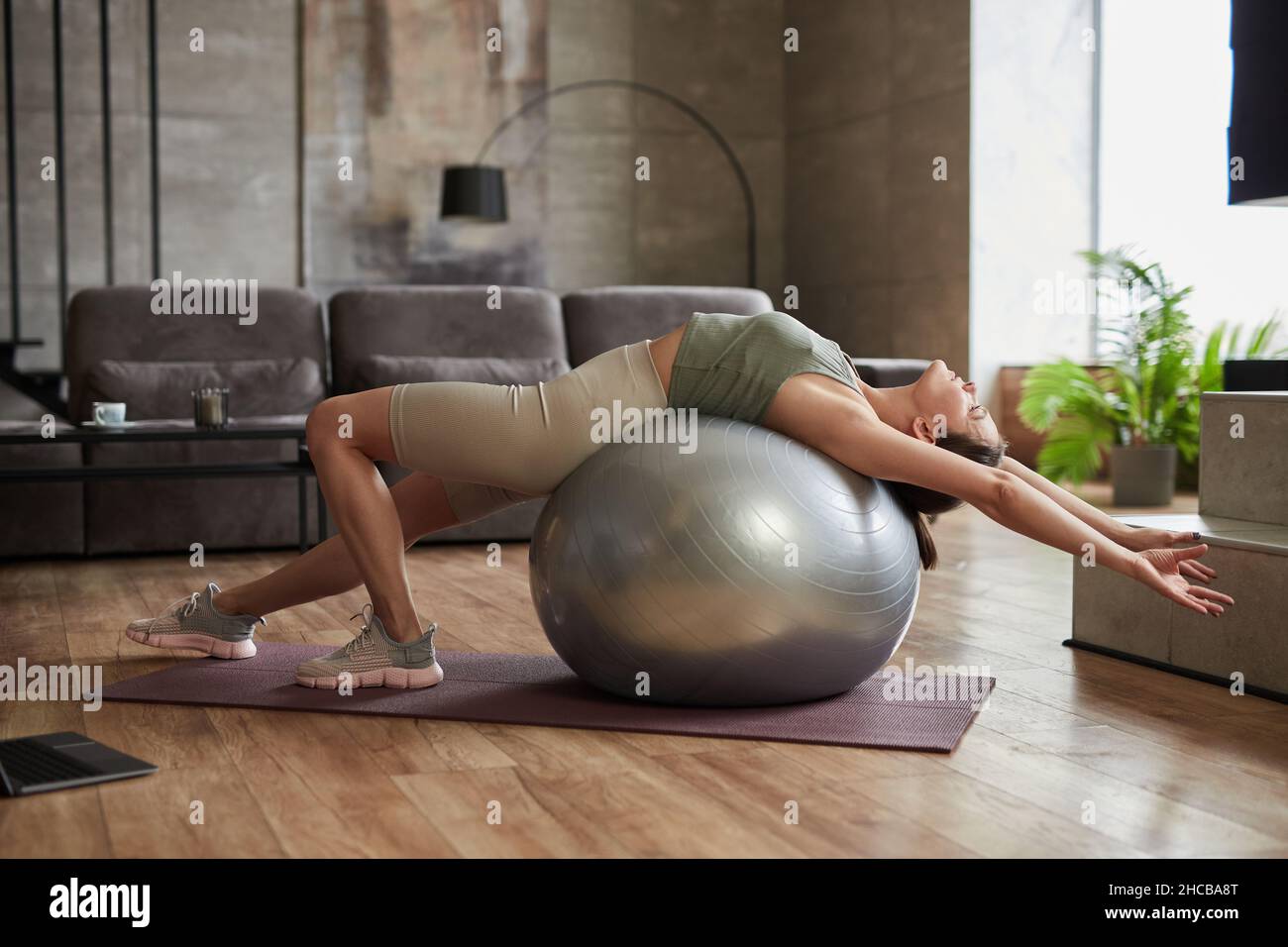 Mujer joven tumbada en la pelota de fitness y haciendo ejercicios de estiramiento durante el entrenamiento en casa Foto de stock