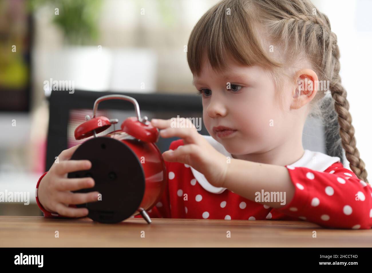 Chica pequeña con colas mirando el reloj despertador rojo en la mesa Foto de stock