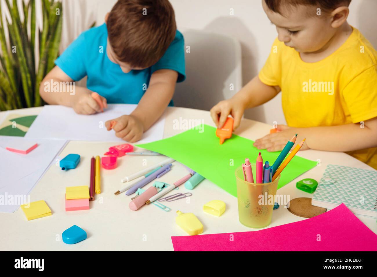 Felices niños con camisas amarillas y azules haciendo artes y artesanías juntas en su escritorio. Los niños dibujan sobre papel coloreado. Los niños juegan en casa. Foto de stock