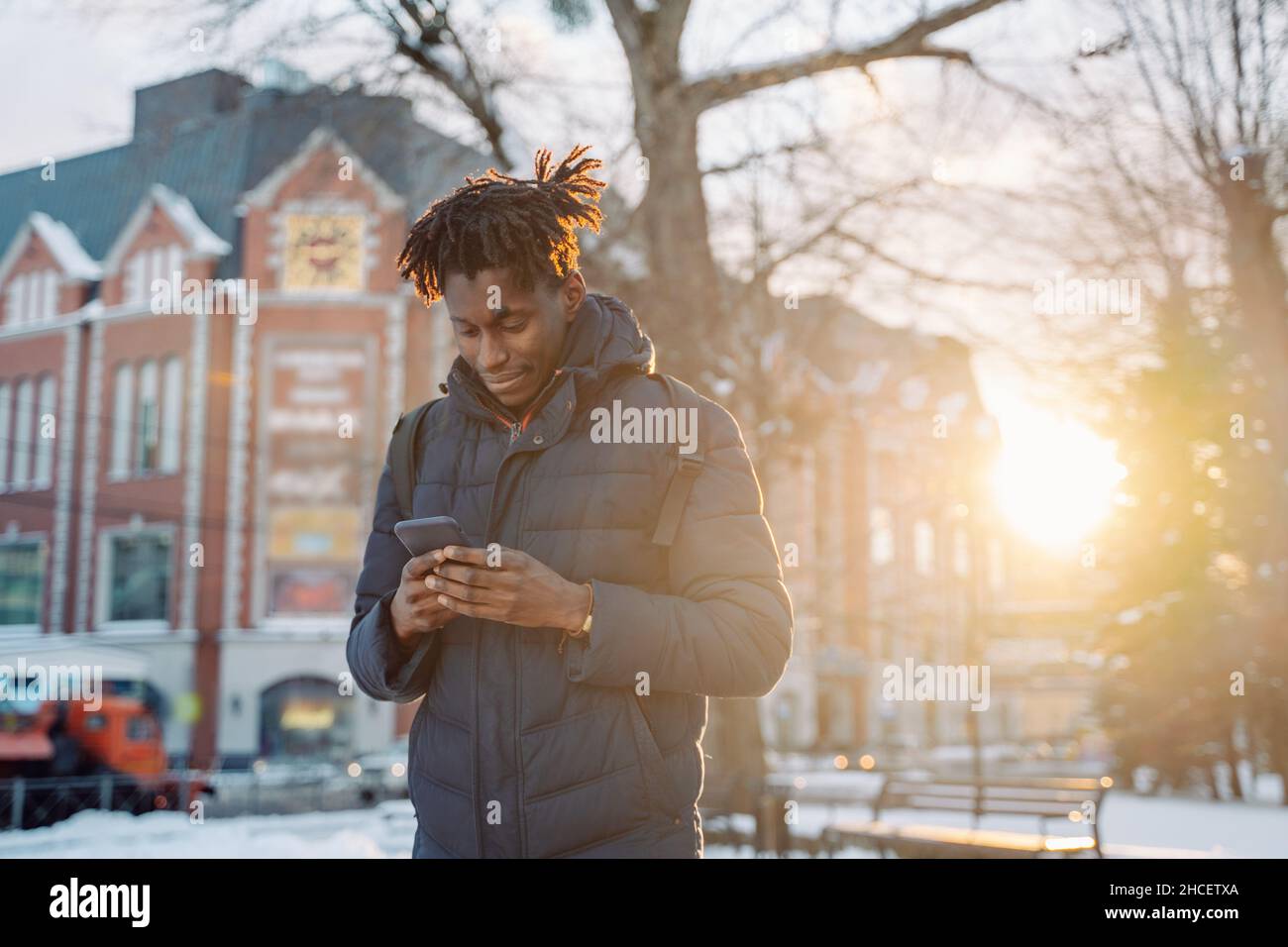 Un joven africano que lleva chaqueta y mochila usa un smartphone en la calle de la ciudad. Turismo, vacaciones en Europa, comunicaciones. Foto de stock