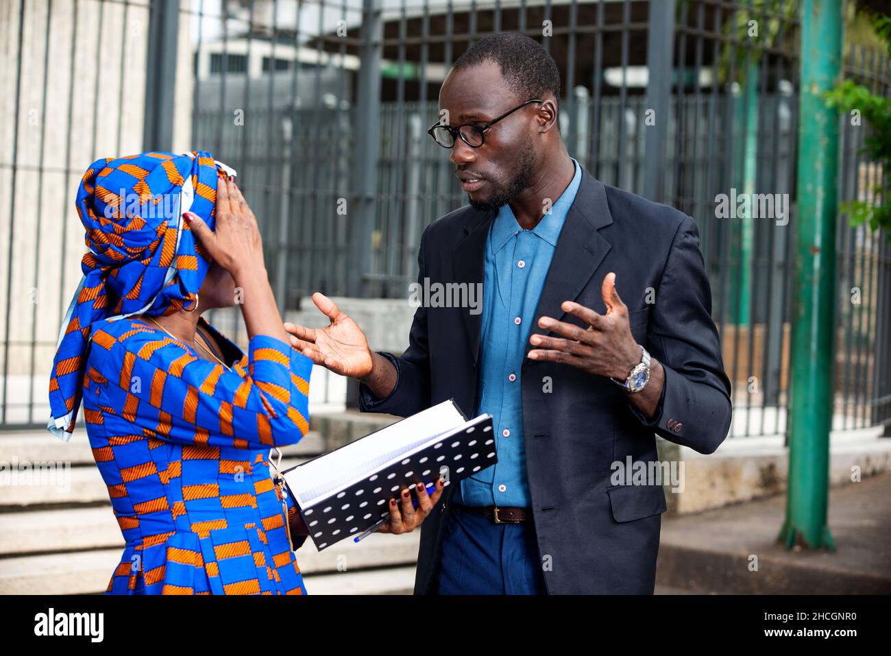 Una hermosa mujer de negocios africana madura con vestido tradicional de pie al aire libre mostrando un documento a su pareja mientras que la última le reprocha. Foto de stock