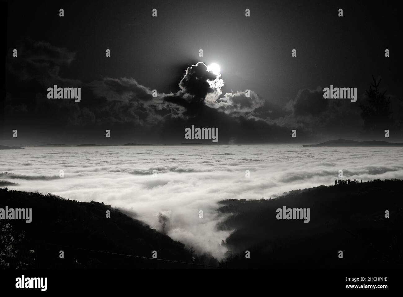 Paisaje rural de montaña por la noche. La Luna que se levanta de las nubes ilumina el mar de niebla en la llanura. Foto de stock