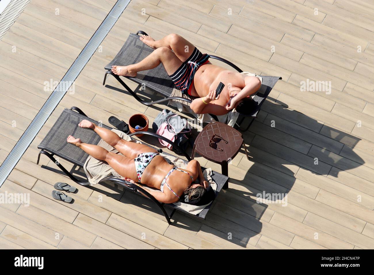 Una pareja tumbada en las tumbonas y tomando el sol cerca de la piscina, vista superior. Mujer relajarse en traje de baño y hombre en los calzones con un smartphone en la playa Foto de stock