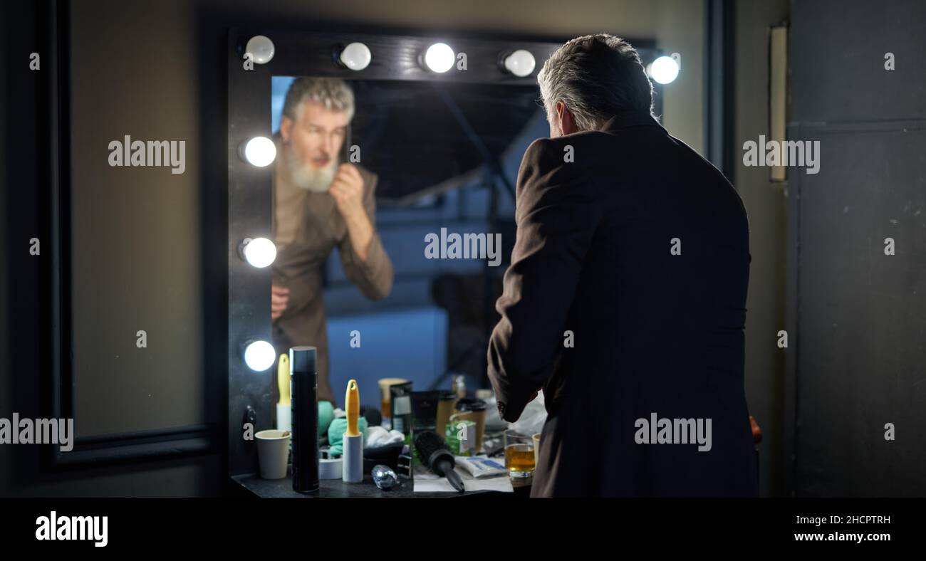Guapo hombre de edad media de pelo gris que lleva un elegante traje mirando a sí mismo en el espejo mientras se prepara para el estudio photohoot Foto de stock