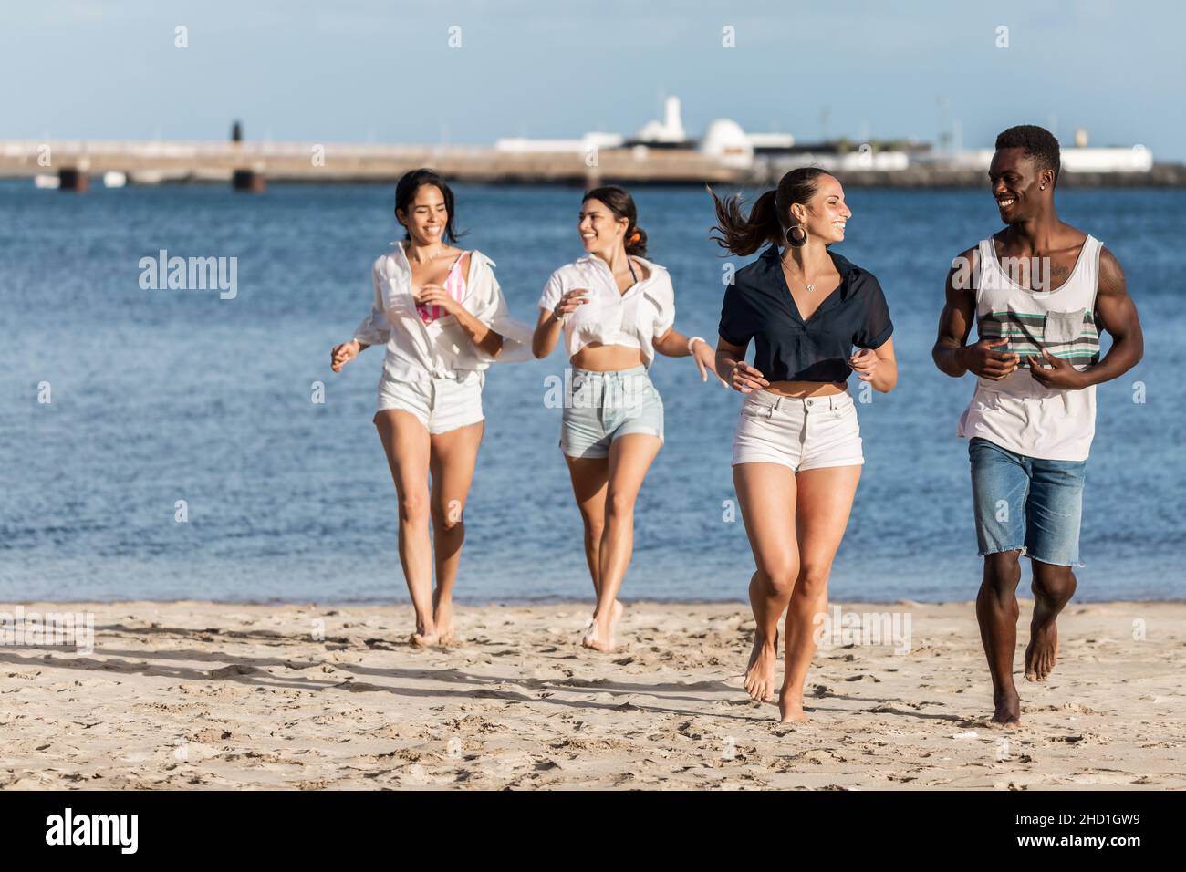 Cuerpo entero descalzo hombre y mujer diversos sonriendo y mirando unos a otros mientras corres en la orilla del mar cerca de las amigas en fin de semana en Lanzarote islan Foto de stock