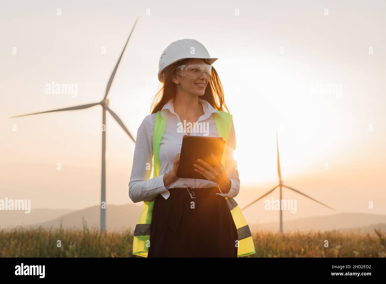 Hermosa mujer caucásica en casco blanco trabajando con tableta digital en la granja de energía renovable. Inspector femenino que controla el funcionamiento de los aerogeneradores al aire libre. Foto de stock
