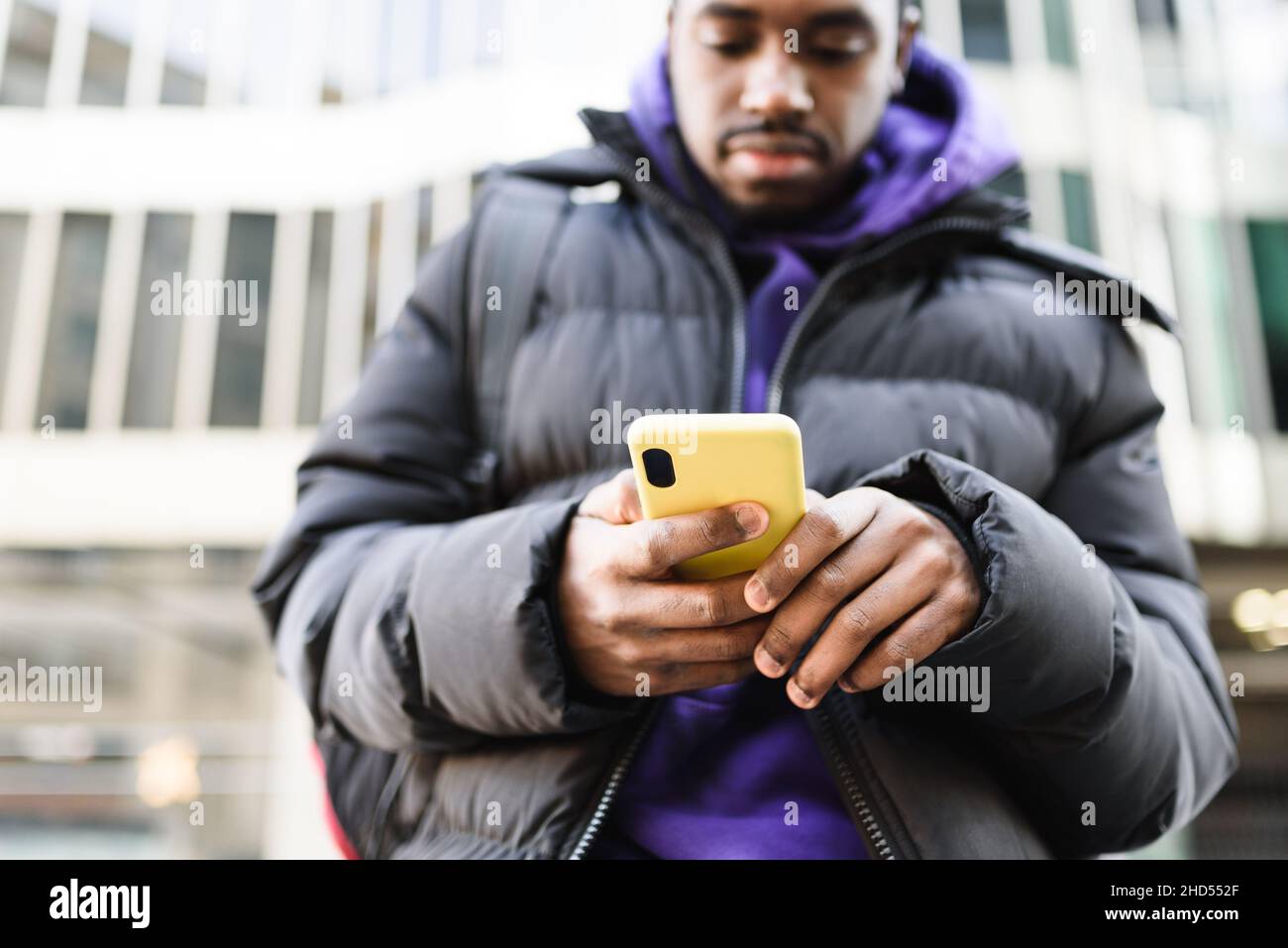 Enfoque suave de macho afroamericano en ropa exterior de navegación celular con caja amarilla mientras se encuentra en la calle de la ciudad moderna Foto de stock