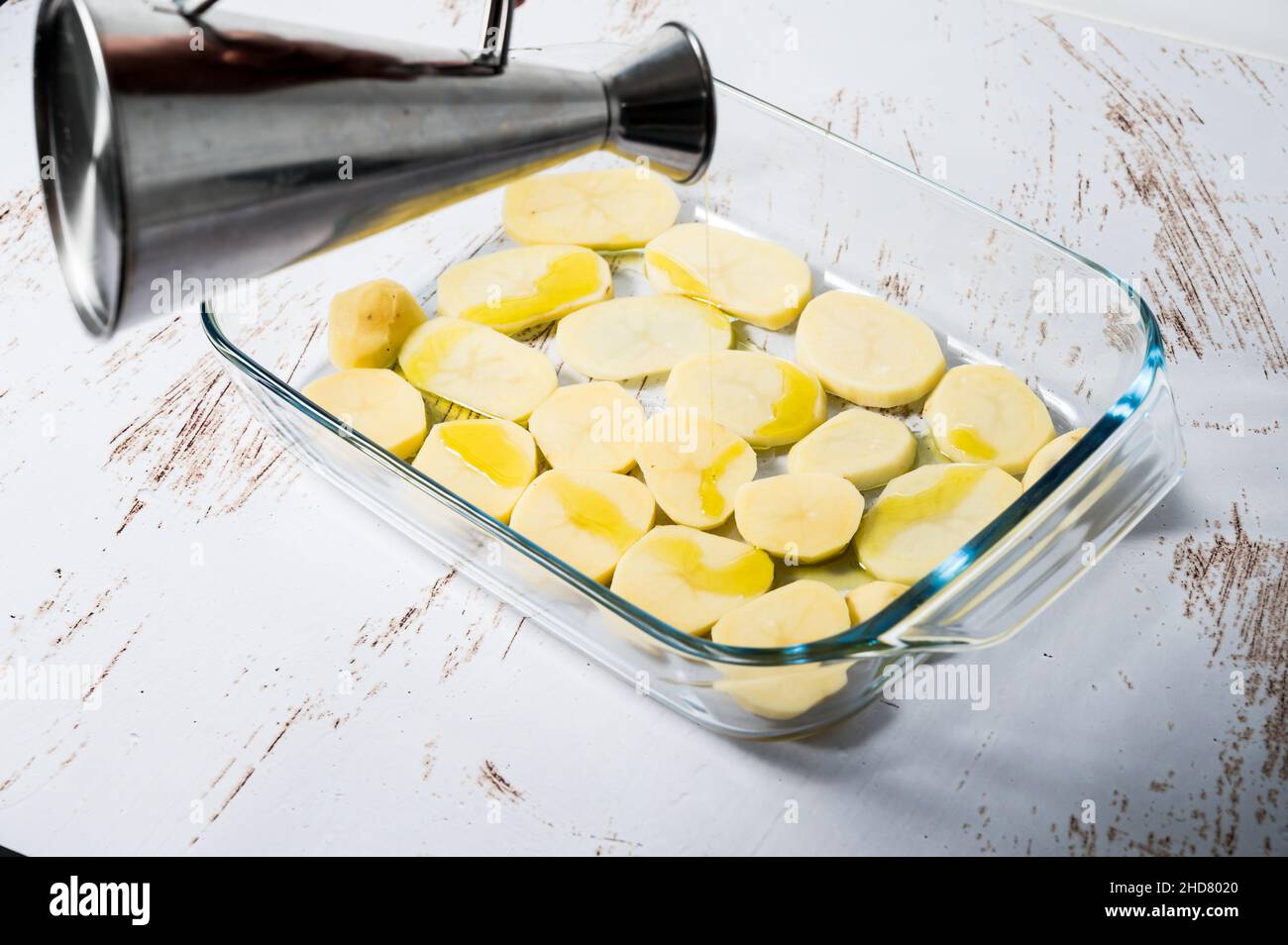 Persona irreconocible que añade aceite a una cama de patatas en un recipiente de cristal para preparar un cava de pollo en el horno. Foto de stock