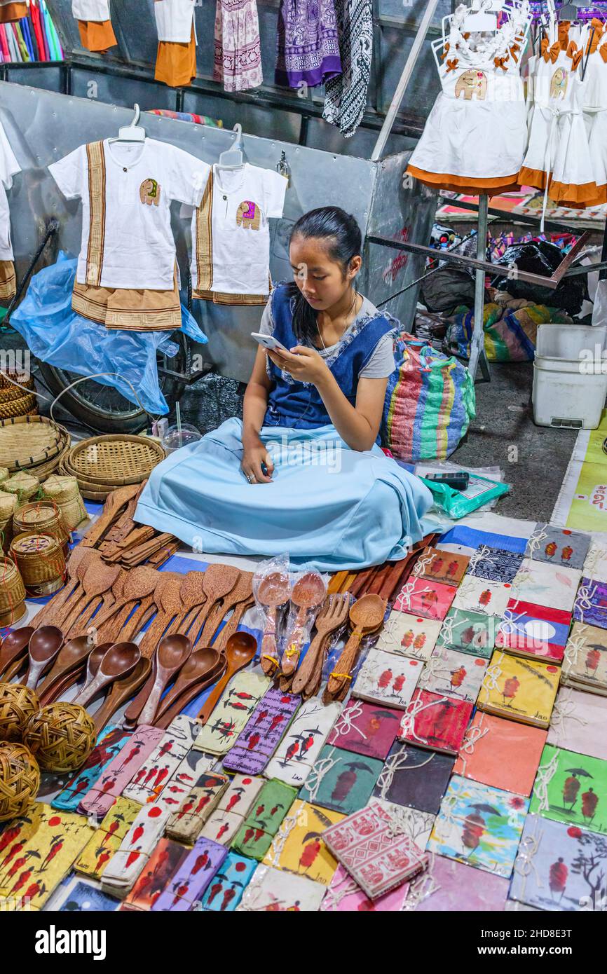 Hermosa joven niña stallholder en un puesto de recuerdo mirando su teléfono móvil en el mercado nocturno de Walking Street, en el centro de Luang Prabang, norte de Laos Foto de stock