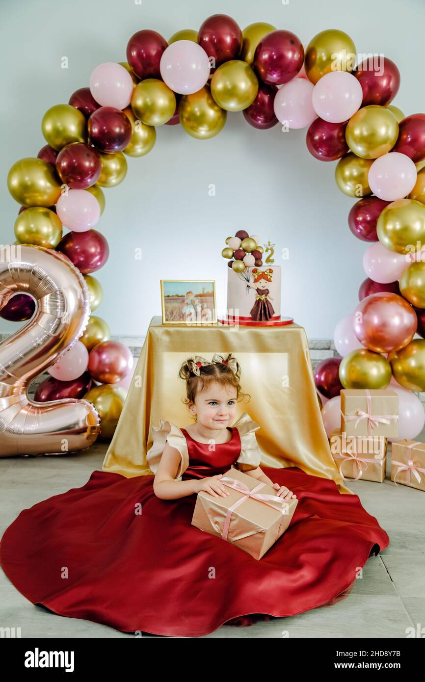 Niña de dos años de edad con un vestido rojo sentado en el suelo frente a  sus decoraciones de cumpleaños y sosteniendo su regalo de cumpleaños.  Nacimiento de una niña encantadora Fotografía
