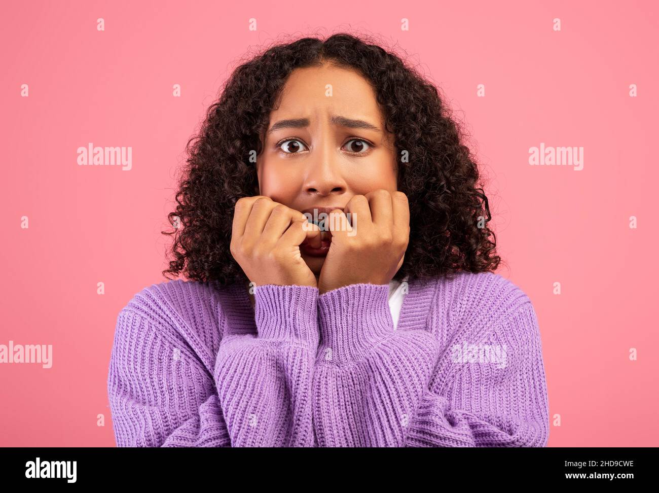 Joven mujer negra que se siente asustada, mordiendo uñas en pánico sobre el fondo rosa del estudio. Concepto de emociones humanas negativas Foto de stock