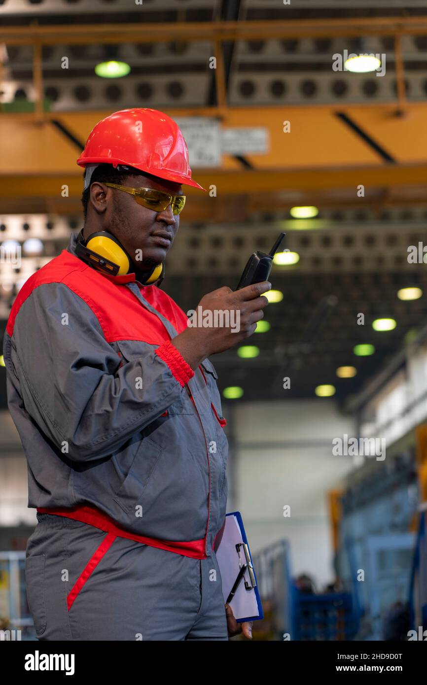 Trabajador afroamericano en ropa de trabajo de protección Hablando sobre equipos de comunicación por radio en una fábrica. Retrato del trabajador industrial negro. Foto de stock