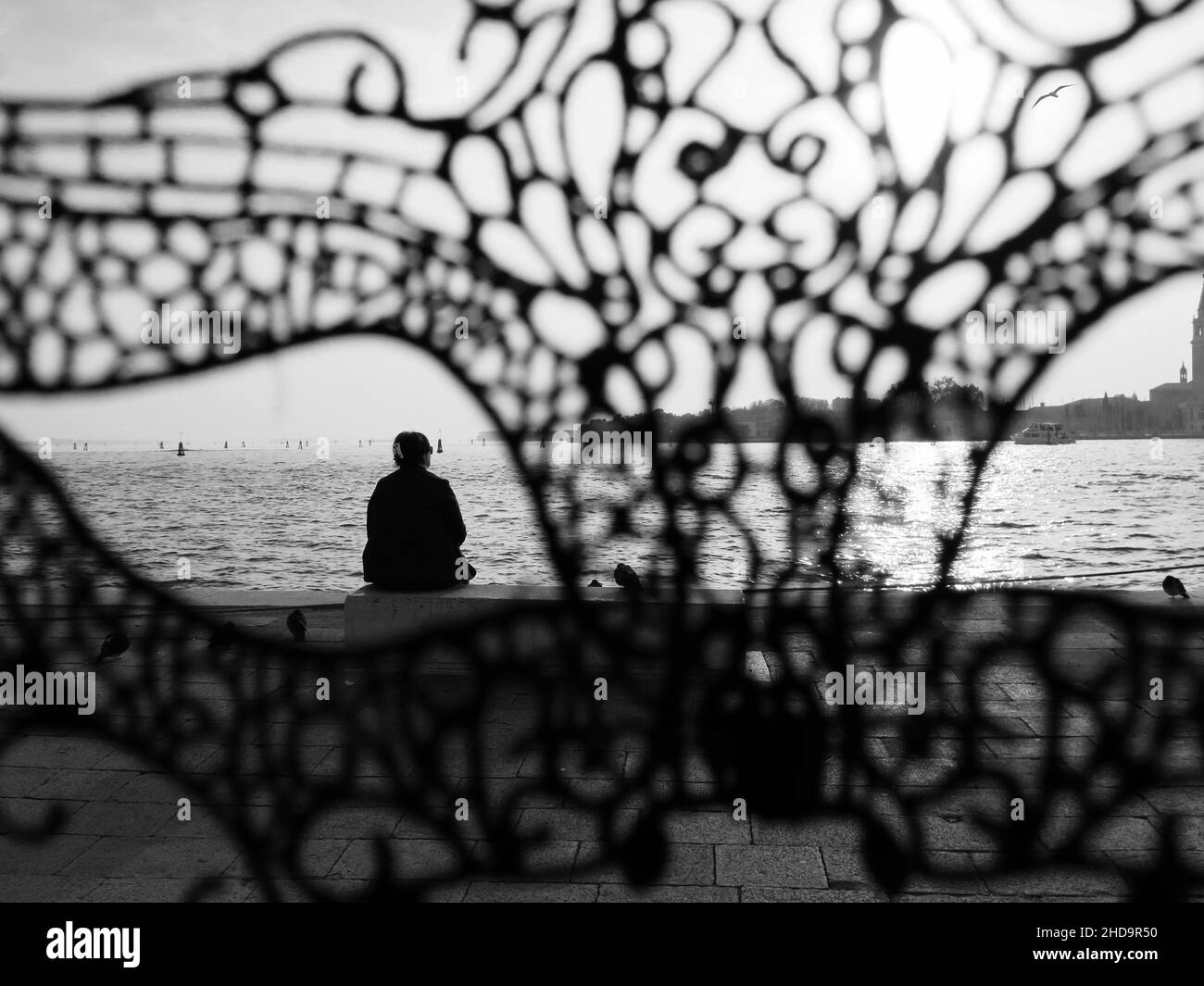 Vista trasera de una persona sentada sola en un banco en la bahía de Venecia, Italia, detrás de una máscara Foto de stock