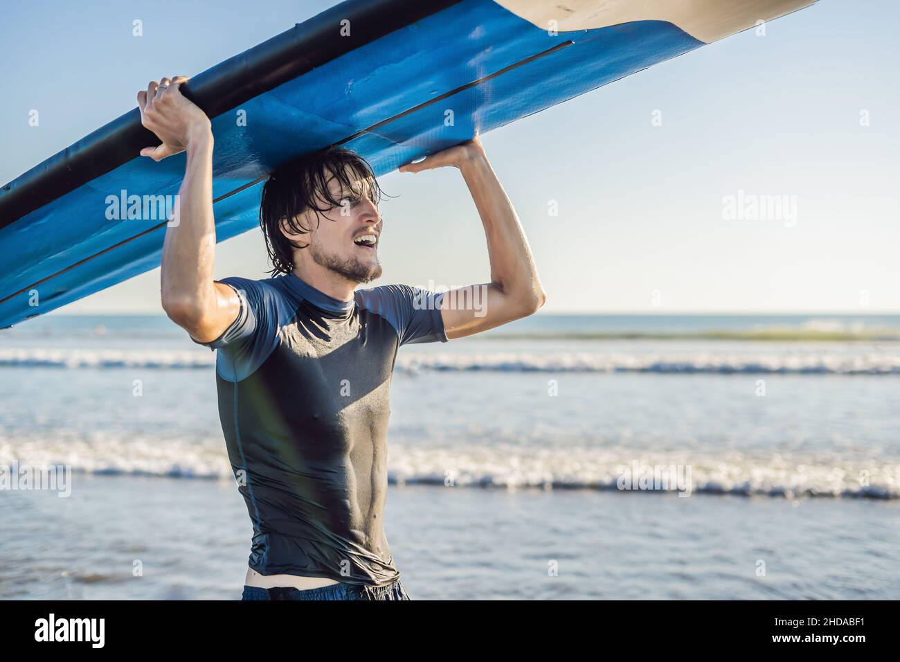 El hombre lleva surf por encima de su cabeza. Cerca de guapo con tablas de surf en la cabeza en la playa. Retrato de hombre con tablas de surf en cabeza y HID Foto de stock