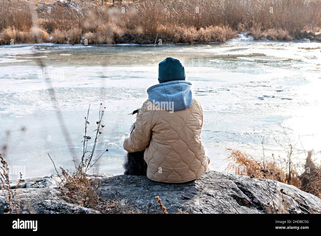 Mujer joven de detrás sentada en perro abrazando de piedra mirando al río cubierto con el primer hielo en otoño o al principio del invierno, color beige neutro Foto de stock