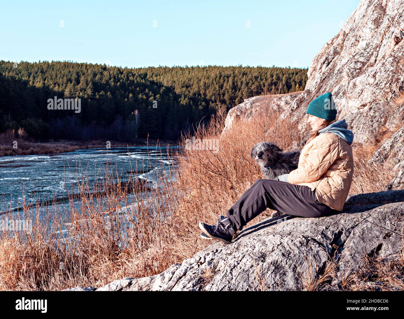 Mujer joven sentada en un perro abrazado de piedra mirando al río cubierto con el primer hielo en otoño o a principios de invierno, colores beige neutros, tonos tierra Foto de stock