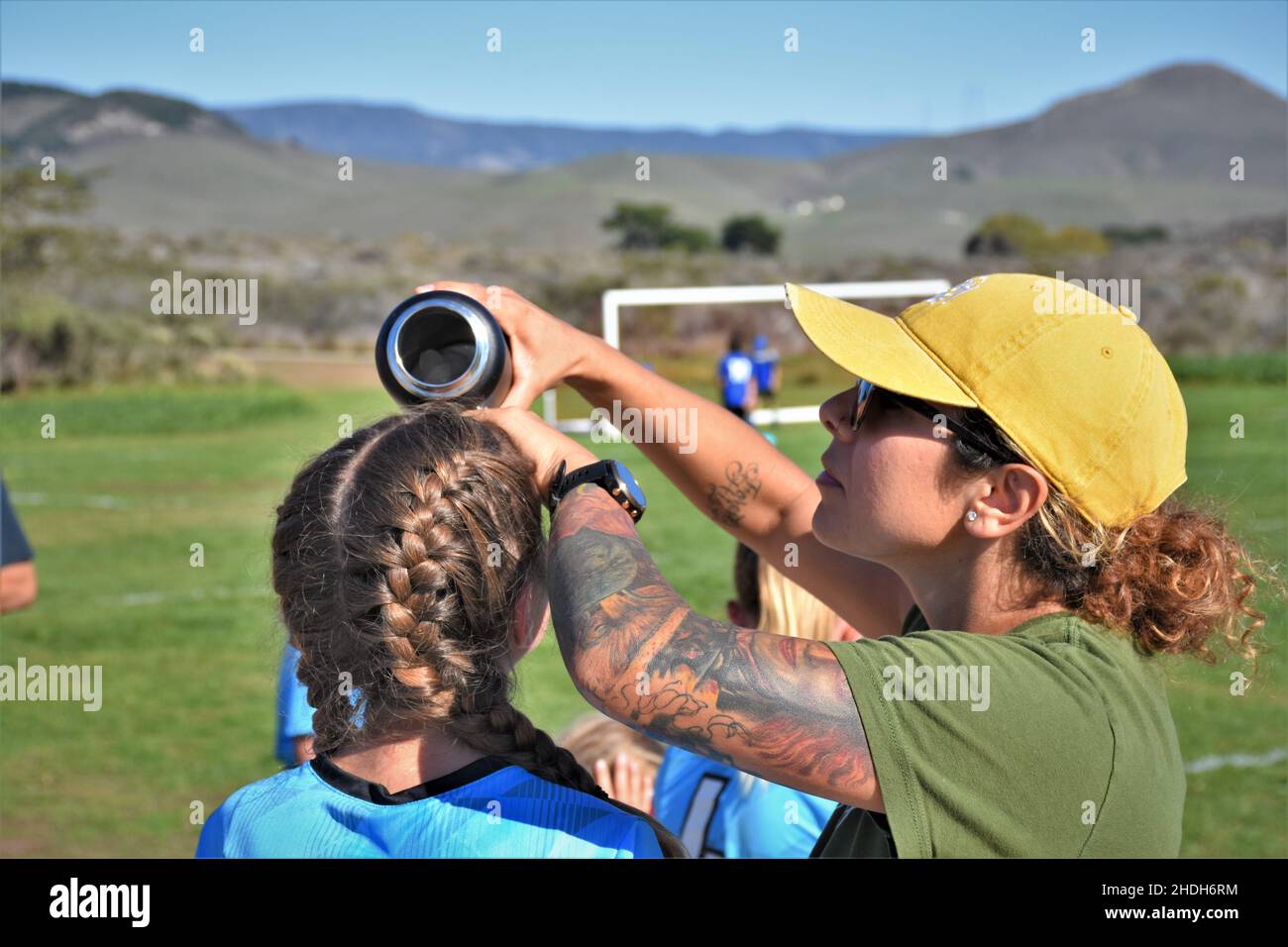 Mamá refrescándose de su chica en la mitad de un partido de fútbol de 14 años de edad en el calor de 95 grados del sur de California con trenzas y tatuajes, gorra Foto de stock
