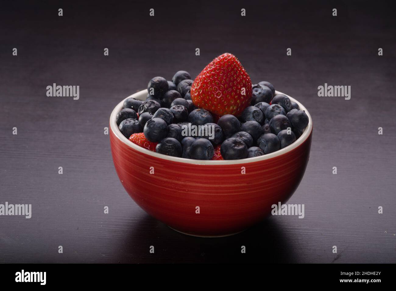 Rojo cuenco lleno de arándanos y fresas sobre mesa negra. Colorida variedad de frutas que abretan la boca esperando a ser consumidas. Foto de stock