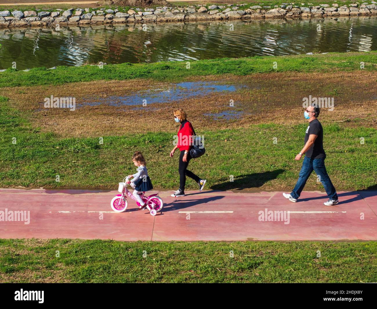 Fuengirola, Málaga, España. 02,06,2021 Un par de abuelos con máscaras faciales recorren la bicicleta de su nieta a través de un parque en un día soleado Foto de stock