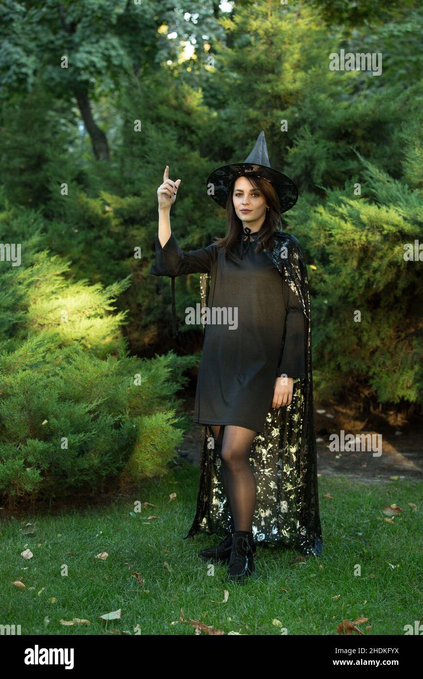 Retrato de una joven atractiva con un vestido negro con una capa sobre un fondo de bosque verde. La chica está usando un sombrero negro puntiagudo con ancho Foto de stock