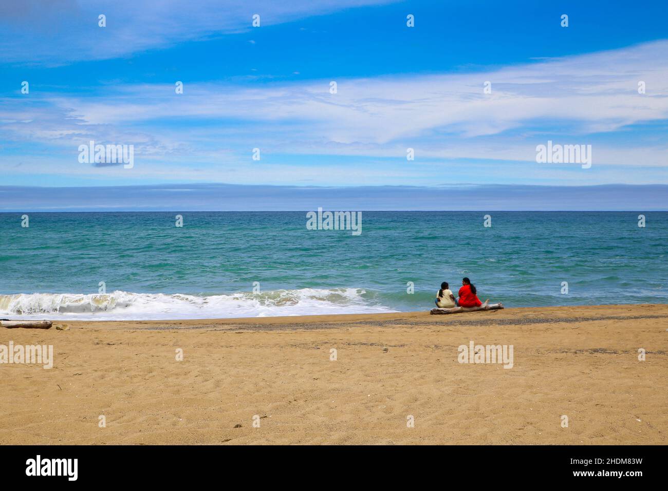 Una madre y una hija se sientan en un registro y observan las olas Adelante. Vacaciones de verano junto a la playa en la Isla del Norte, Nueva Zelanda Foto de stock