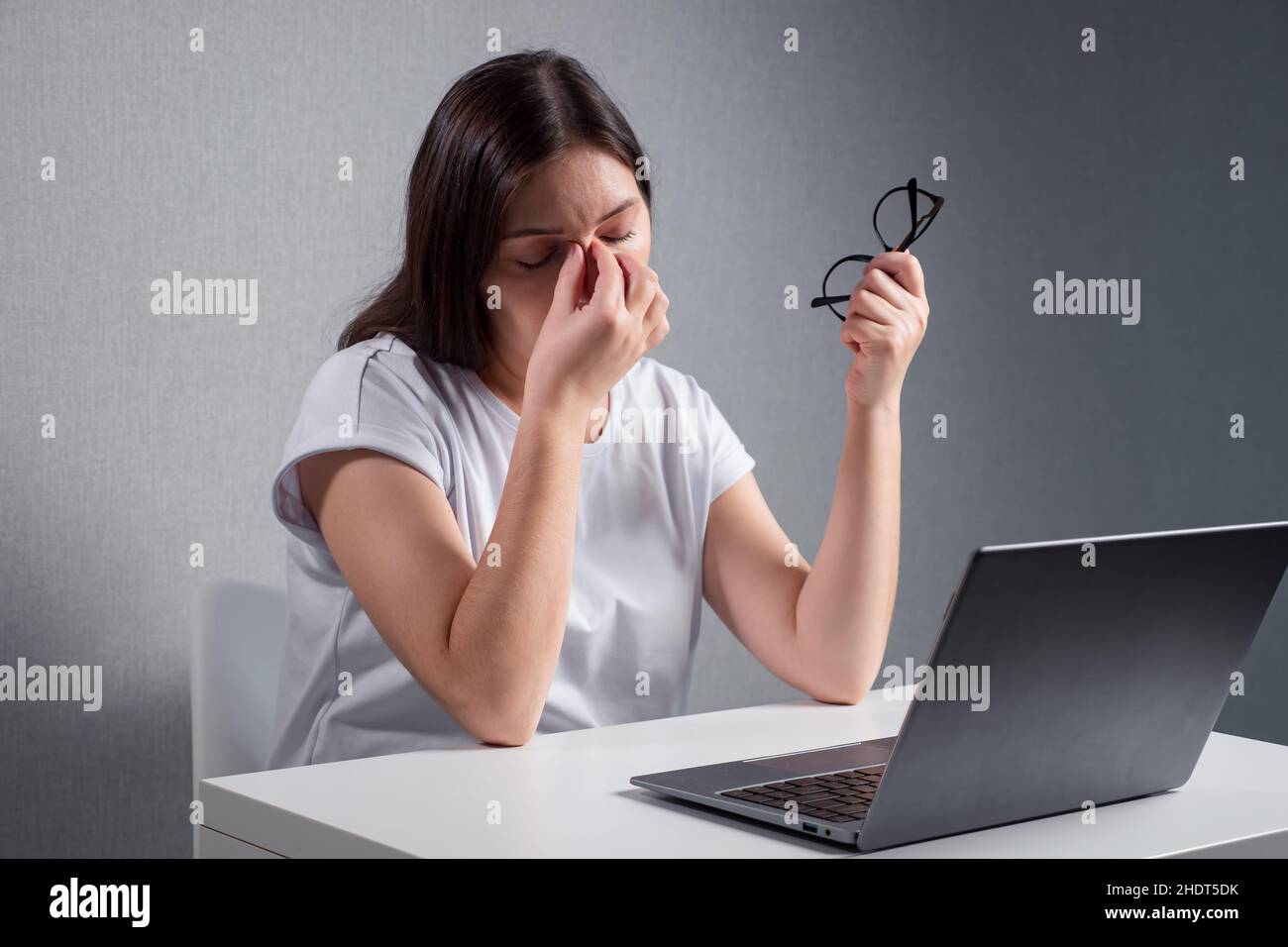 mujer joven masajeando el puente de la nariz, sosteniendo las gafas en otra mano, sentada delante de un ordenador portátil Foto de stock