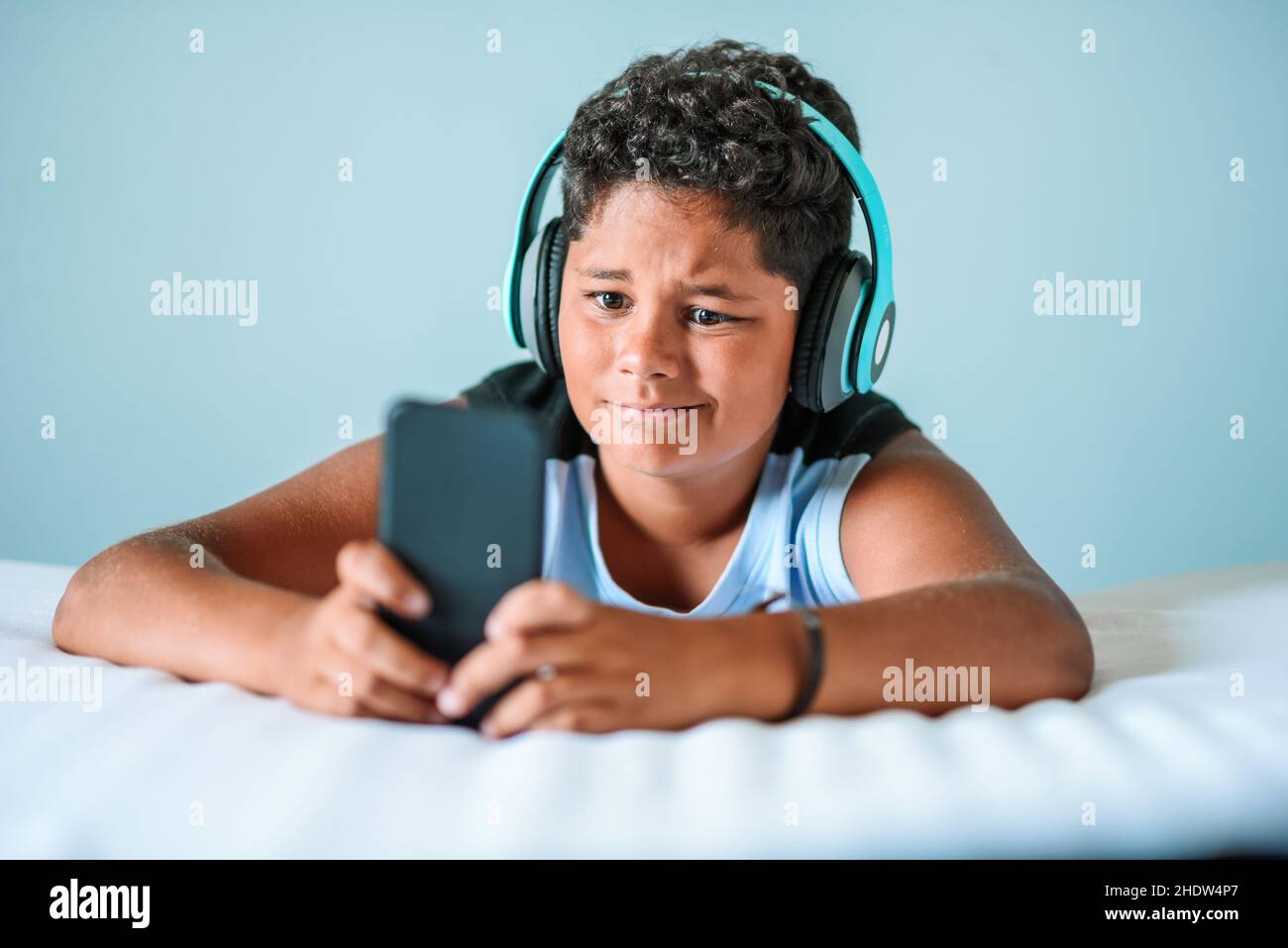 Un chico étnico decepcionado con pelo rizado oscuro en unos modernos auriculares que juegan a videojuegos en un smartphone mientras se encuentra en una cómoda cama a la luz del día en casa Foto de stock