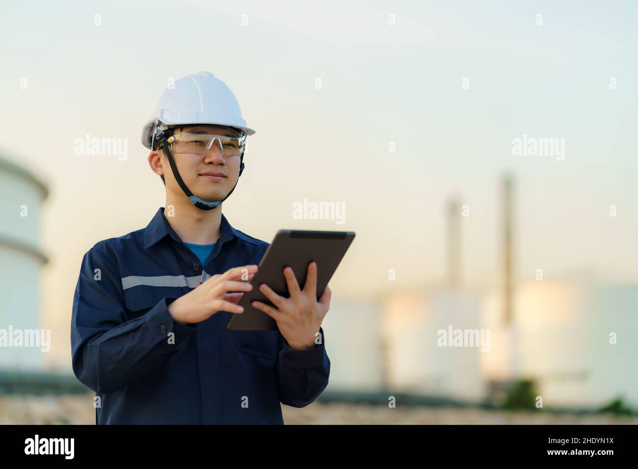 El ingeniero asiático está comprobando el mantenimiento de la fábrica de refinería de petróleo por la noche a través de tabletas digitales. Foto de stock