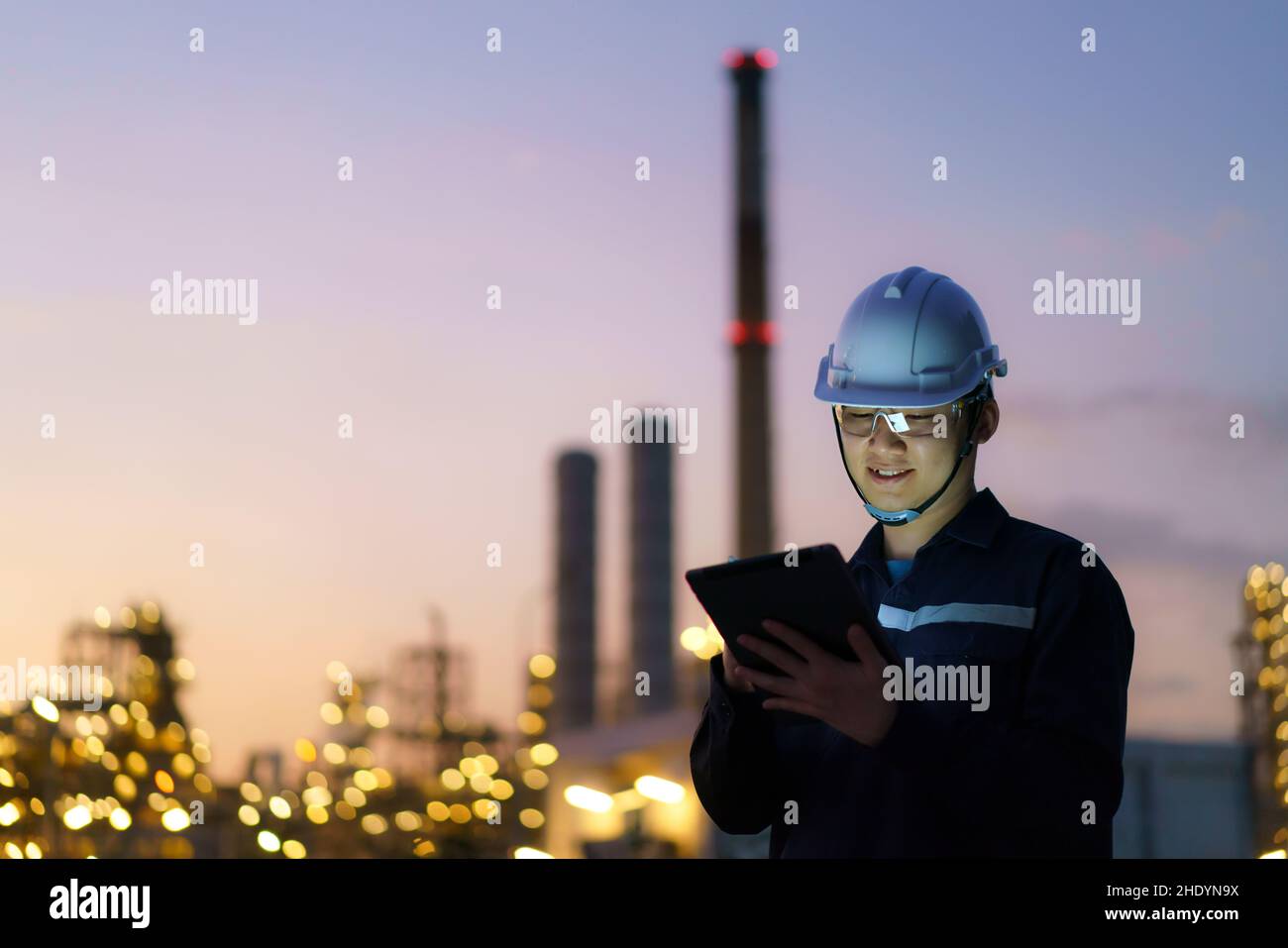 Ingeniero asiático que comprueba el mantenimiento de la fábrica de refinería de petróleo por la noche a través de tabletas digitales. Foto de stock